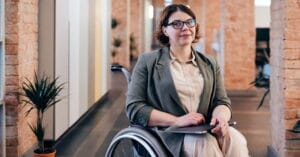 A woman holding a laptop, sitting in a wheelchair in an open plan office