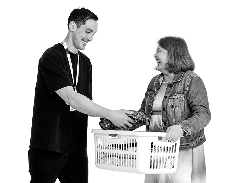 A support worker and participant laughing together while carrying a laundry basket during in home support