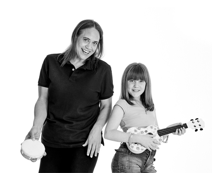 A woman holding a tambourine standing next to a girl holding a ukelele. Both smile at the camera.