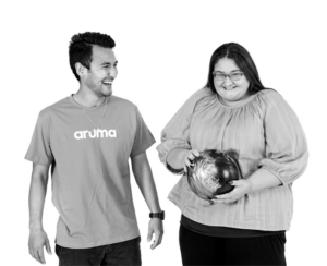A young woman holding a bowling ball smiles alongside an Aruma support worker during a community participation outing.