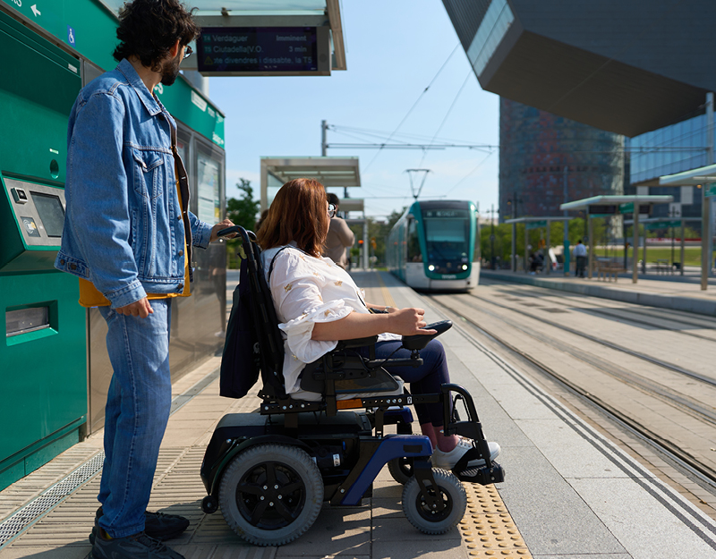 A woman in a power wheelchair and a support worker buying a ticket at a tram stop during capacity building support