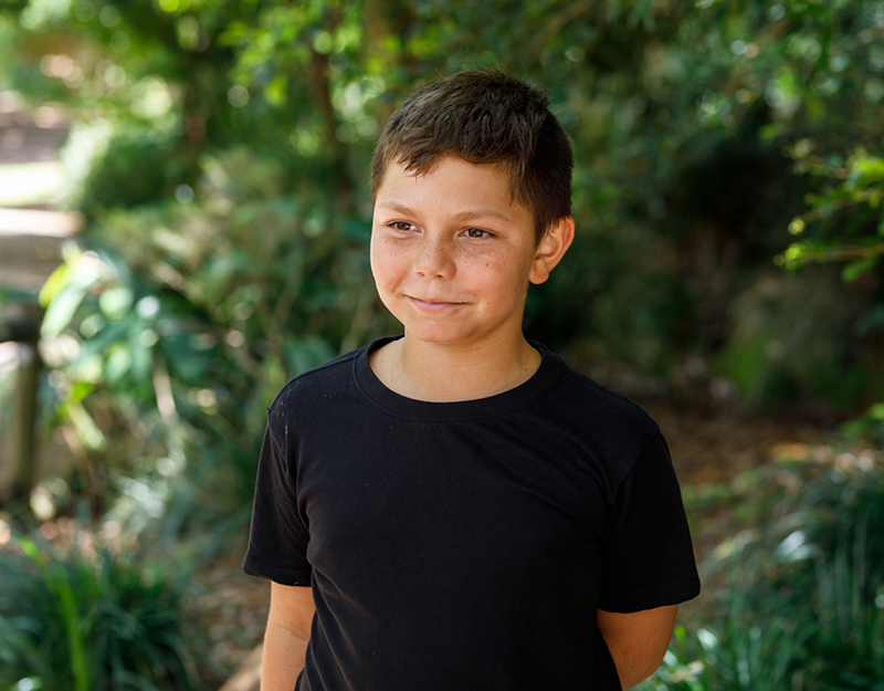 A young boy standing outdoors in a garden setting, smiling gently, representing psychology services for children