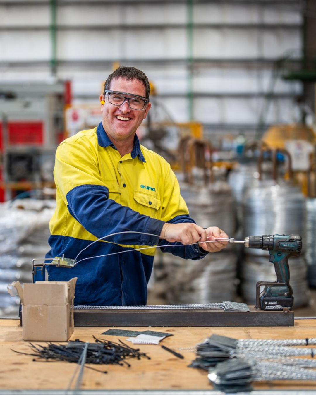 Craig smiling while using a drill at Aruma Manufacturing, wearing safety glasses and high-vis workwear.