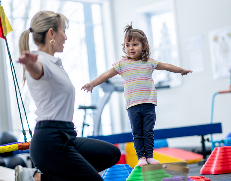 A young girl balancing on colourful stepping stones with arms outstretched while an occupational therapist guides her