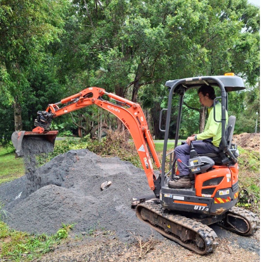 A man in a hi-vis shirt operates a small orange Kubota excavator, tipping a bucket of gravel onto a pile. He is on a driveway surrounded by green trees.