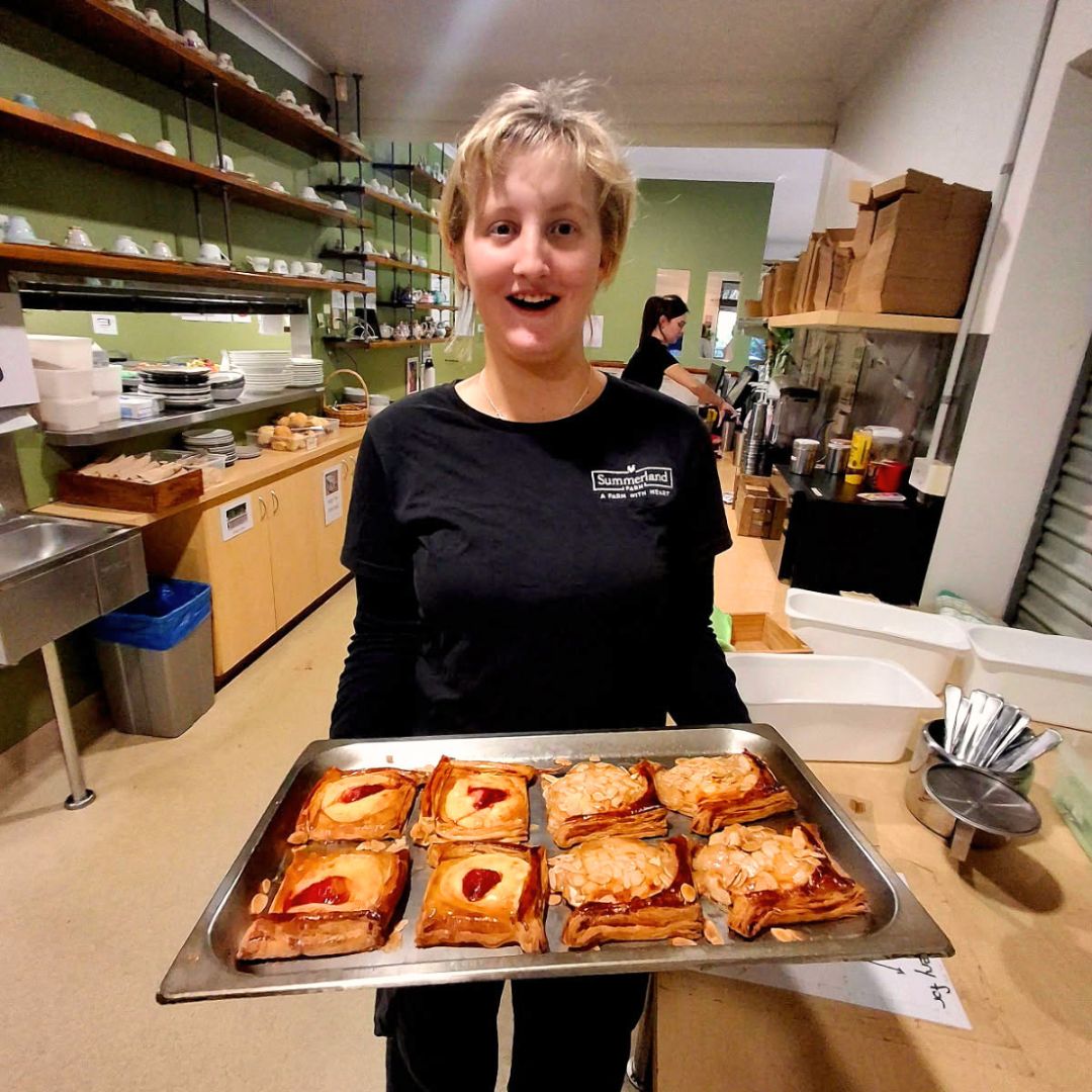A young woman with short blonde hair in a black Summerland Farm t-shirt smiles widely as she holds a tray of freshly baked almond and jam danishes in a cafe kitchen.