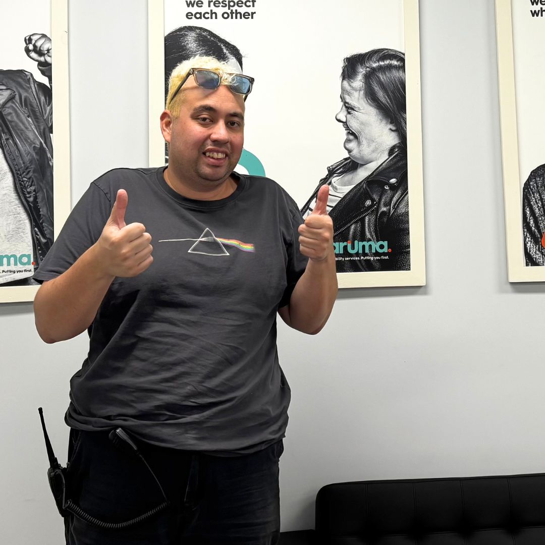A young man in a dark t-shirt gives two thumbs up and smiles, standing in front of framed black-and-white Aruma posters on a pale wall.