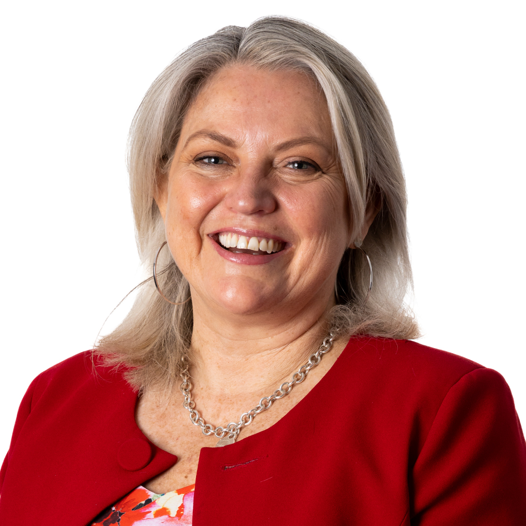 Lylea McMahon, Chief Operating Officer, smiling brightly. She is wearing a red top and silver chain necklace with hoop earrings, with shoulder-length silver-grey hair.