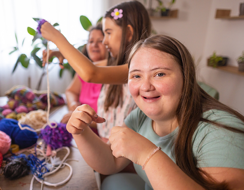 Targeted Care Packages: A young woman smiles while doing a craft activity with colourful yarn. Two other people work on crafts in the background in a bright, homely room.