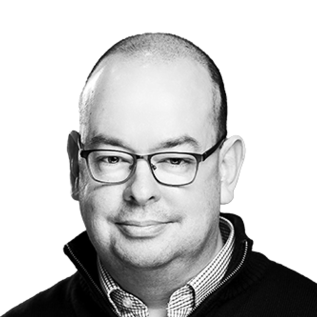 Black and white portrait of Cain Beckett, Director, with a relaxed smile. He is wearing glasses and a dark jacket over a checked shirt, with short cropped hair.