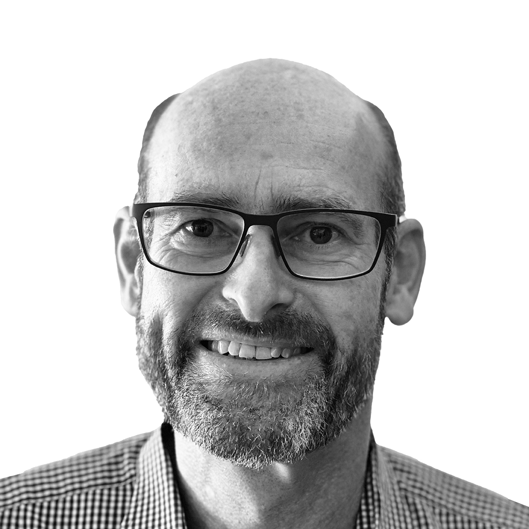 Black and white portrait of Dr Joost de Kock, Director, smiling. He is wearing glasses and a checked shirt, with a beard and shaved head.
