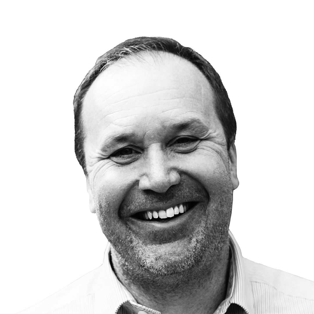  Black and white portrait of Phil Wade, Director, smiling broadly. He has short hair and stubble, and is wearing a light striped shirt.