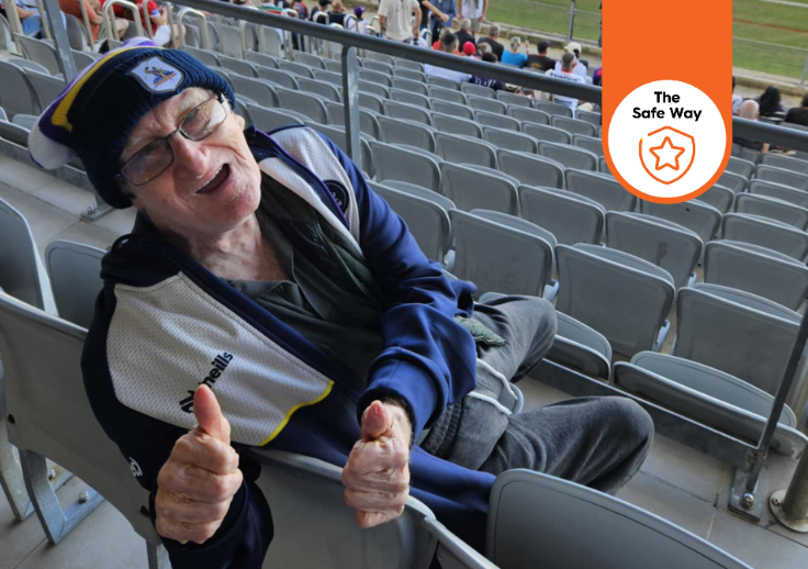 George leans over the back of the seat with the stadium in the background, he's giving the camera two thumbs up