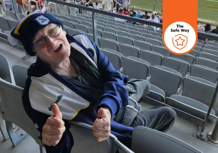 George leans over the back of the seat with the stadium in the background, he's giving the camera two thumbs up