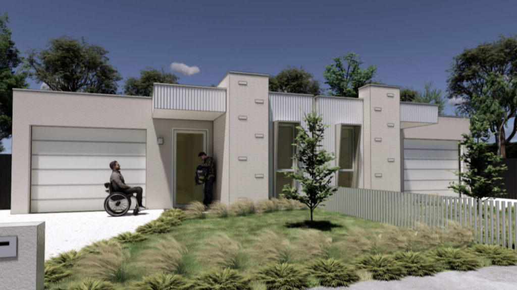Front exterior of one of the Ringwood East SDA homes, showing a wheelchair user and support worker at the accessible front entrance, with a white and grey rendered facade, garage, landscaped native garden and front fence.