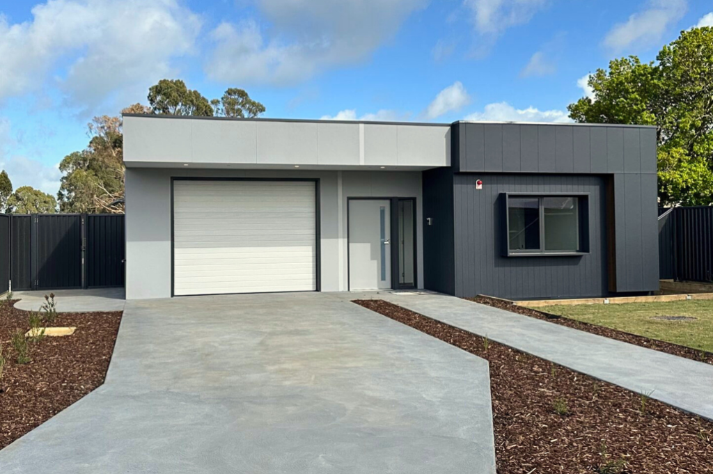 Front exterior of one of the Long Gully SDA homes, showing a single-storey modern home with dark grey and white cladding, garage, front door, landscaped garden beds with mulch and a grassed front yard.