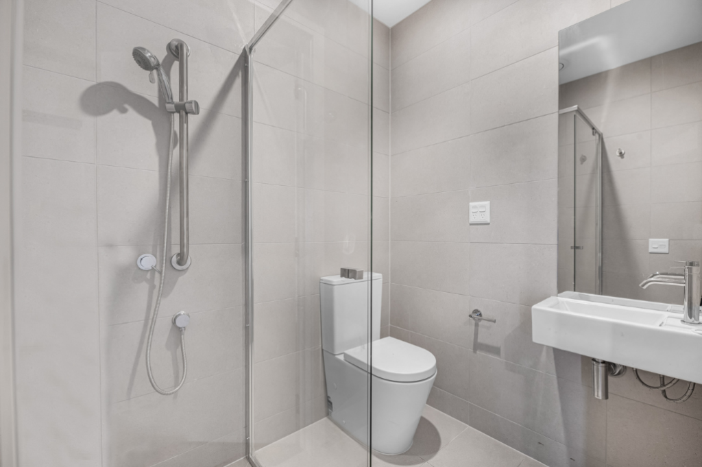 Bathroom inside one of the Long Gully SDA homes, showing a walk-in shower with glass screen, wall-mounted basin with mirror, toilet and tiled flooring.