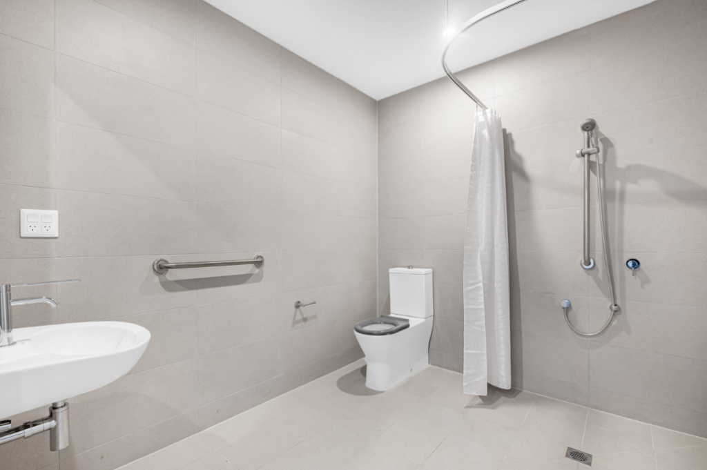 Accessible ensuite inside one of the Long Gully SDA homes, showing a roll-in shower with curtain rail, grab rails, wall-mounted basin, toilet and tiled flooring.