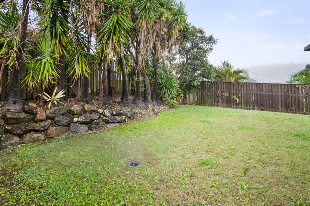 Backyard of the Pacific Pines SIL home, a fully fenced grassed area bordered by established palms and a rock garden.