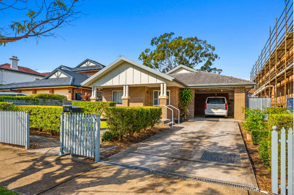 Front exterior of the Strathfield SDA home, showing a single-storey brick home with a gabled entry porch, carport with vehicle, ramp with handrails to the front entry, landscaped hedges and picket fence.