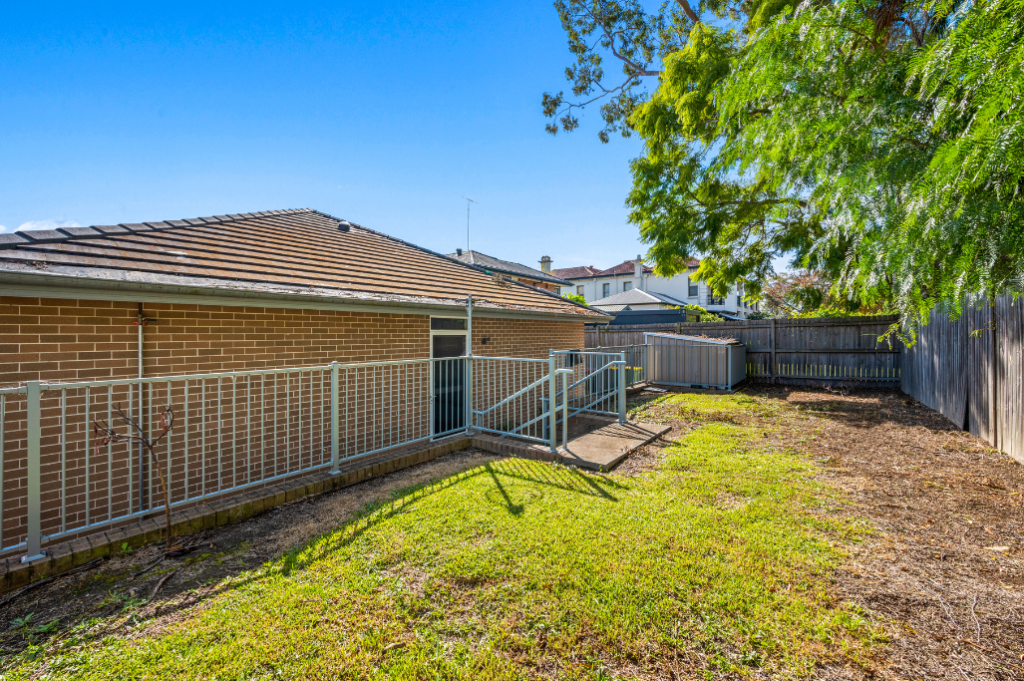 Backyard of the Strathfield SDA home, showing a fully fenced grassed area with mature tree, accessible ramp with handrails from the rear of the home and a garden shed.