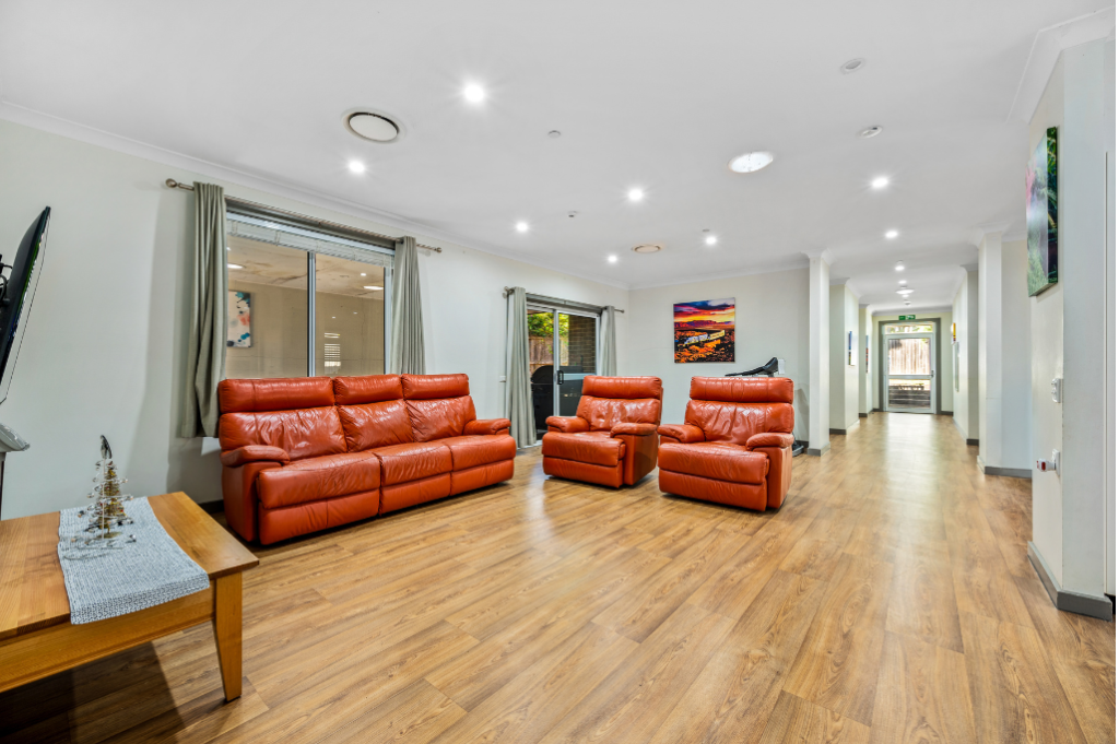 Main living room inside the Strathfield SDA home, showing a leather lounge suite with recliner, wall-mounted TV, coffee table, sliding doors to outdoor areas, artwork on walls and wide hallway leading to bedrooms.