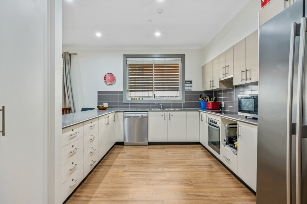 Kitchen inside the Strathfield SDA home, showing white cabinetry, grey tiled splashback, stainless steel dishwasher, oven, large fridge and timber-look flooring.