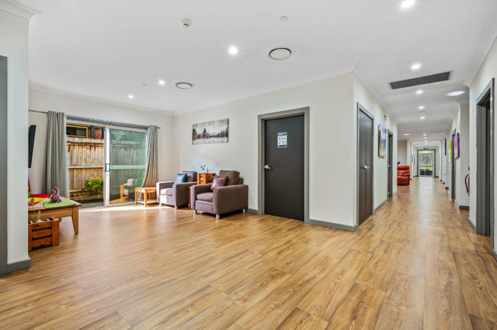 Second living area inside the Strathfield SDA home, showing armchairs, sliding door to backyard, artwork on walls, wide hallway with bedroom doors and timber-look flooring.