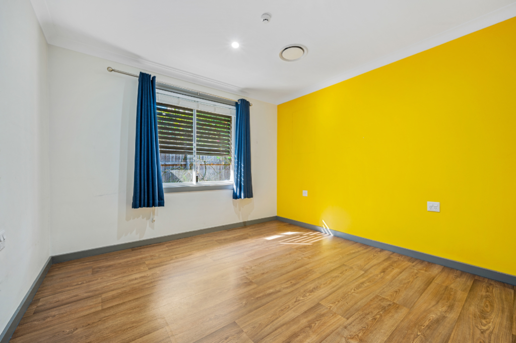 Vacant bedroom inside the Strathfield SDA home, showing a bright room with a yellow feature wall, timber-look flooring, window with blue curtains and natural light.