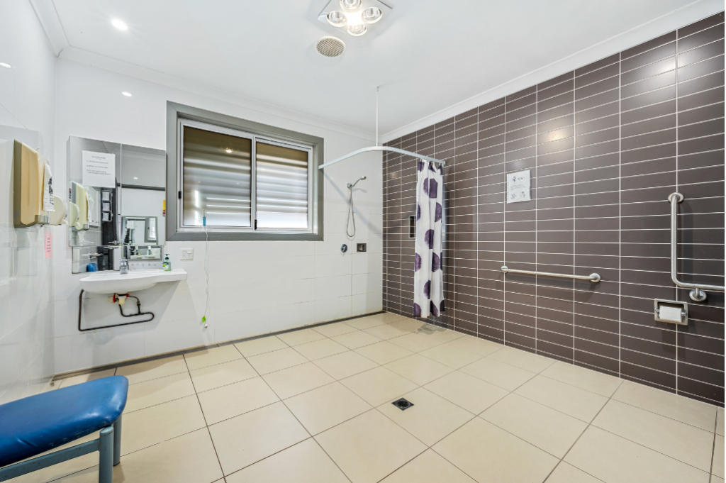 Accessible bathroom inside the Strathfield SDA home, showing a roll-in shower with curtain rail and grab rails, wall-mounted basin with mirror, shower bench, tiled flooring and window.