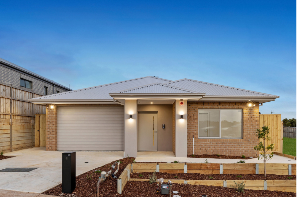 Front exterior of the Sunbury SDA home at dusk, showing a single-storey brick home with a covered entry porch, garage, front windows with exterior lighting, landscaped garden beds with mulch and new plantings.