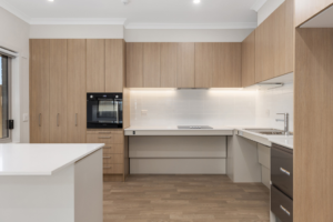 Kitchen inside the Sunbury SDA home, showing timber cabinetry, white stone benchtops with adjustable height section, built-in oven, induction cooktop, under-cabinet lighting and timber-look flooring.