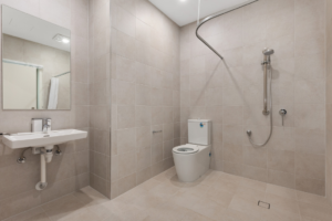 Accessible ensuite bathroom inside the Sunbury SDA home, showing a roll-in shower with curved curtain rail, wall-mounted basin with mirror, toilet and tiled flooring.