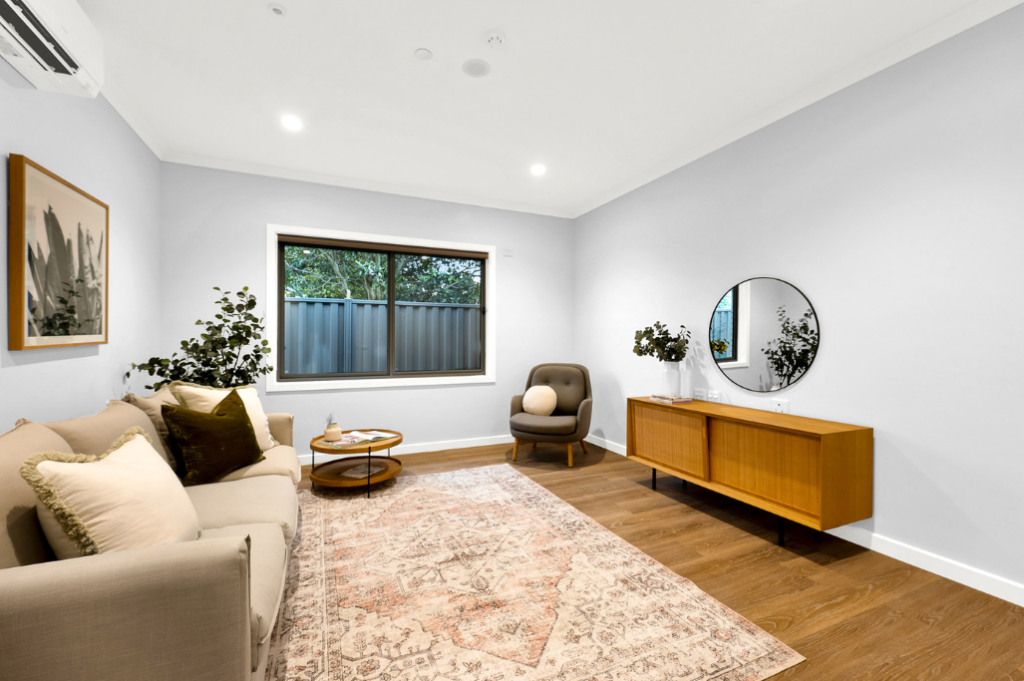 Second living room inside the Sunbury SDA home, showing a sofa, armchair, timber sideboard, artwork, rug and a window looking out to the backyard.