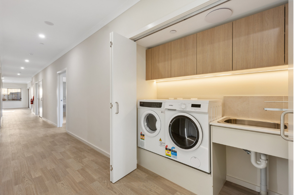 Laundry inside the Sunbury SDA home, showing a front-loading washing machine and dryer, accessible height tub, overhead timber cabinetry with under-cabinet lighting, and a wide hallway visible beyond.