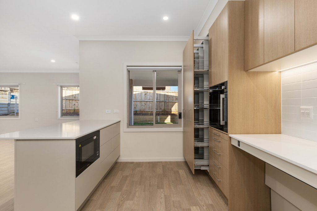 Kitchen inside the Sunbury SDA home from a different angle, showing timber cabinetry, stone benchtops, built-in oven, pull-out pantry storage, island bench and windows looking out to the backyard.