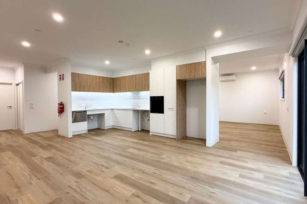 3. Open-plan living, dining and kitchen of the Sale SDA home, showing timber-look flooring, recessed ceiling lights, wall-mounted air conditioning and an accessible kitchen with timber-look cabinetry.
