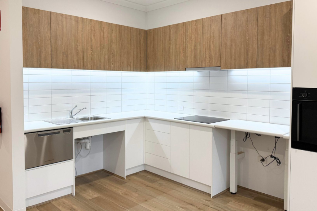 4. Accessible kitchen inside the Sale SDA home, showing an L-shaped layout with white benchtops, tiled splashback, dishwasher, wall oven and timber-look upper cabinetry.