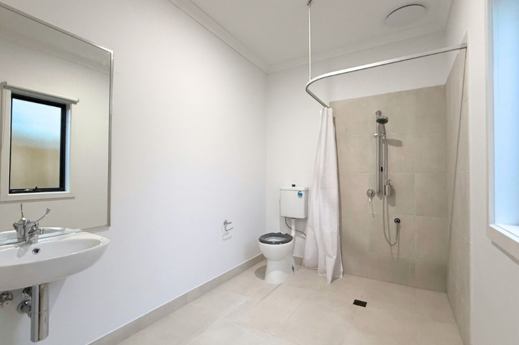 9. Accessible ensuite inside the Sale SDA home, with wall-mounted basin, mirror, accessible toilet and roll-in shower with curtain rail.