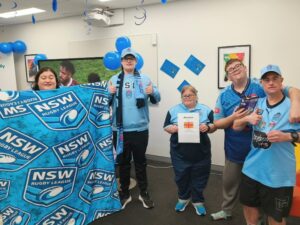 Five Club Aruma Charlestown participants and staff in NSW Blues jerseys and caps, smiling at the camera. One holds a Blues blanket, another holds an NRL Shop voucher. Blue balloons and streamers decorate the room behind them.