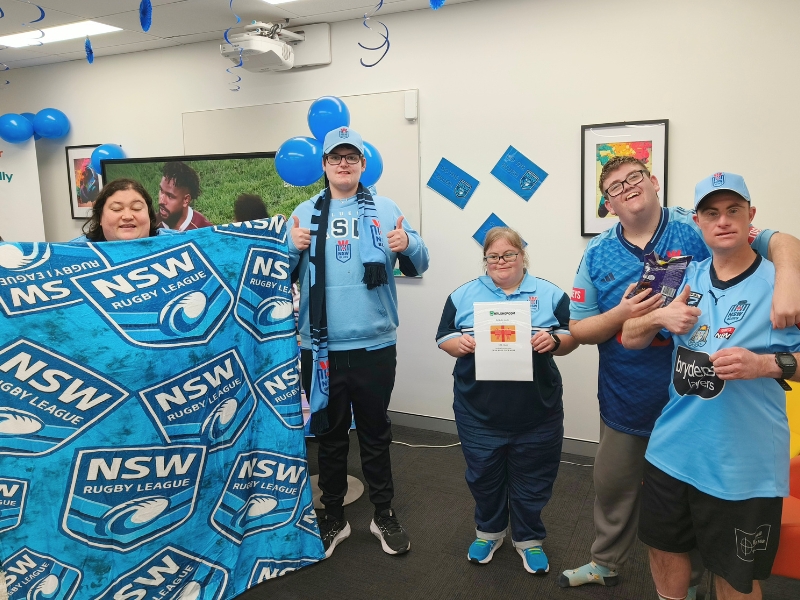 Five Club Aruma Charlestown participants and staff in NSW Blues jerseys and caps, smiling at the camera. One holds a Blues blanket, another holds an NRL Shop voucher. Blue balloons and streamers decorate the room behind them.