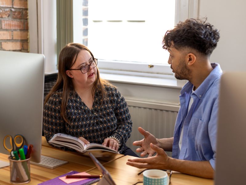 Two people sit at a desk in conversation, with an open notebook between them outlining NDIS changes.