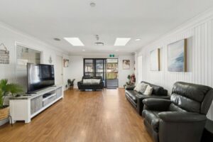 Living room inside the Albion Park SIL home, with timber-look flooring, lounge seating, a large TV, skylights and sliding doors opening to the covered deck.