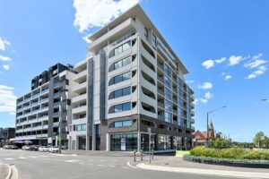 Front exterior of the Luminess Essendon building, a multi-storey modern apartment development with arched balcony detailing, ground-floor retail and street corner location.