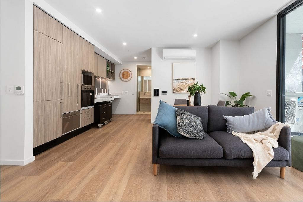 Open-plan living and kitchen area inside the Essendon SDA apartment, showing timber-look flooring, an accessible kitchen with integrated appliances, a lounge and split system air conditioning.