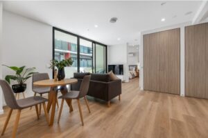 Living and dining area inside the Essendon SDA apartment, showing timber-look flooring, dining setting, lounge and floor-to-ceiling windows opening to the balcony.