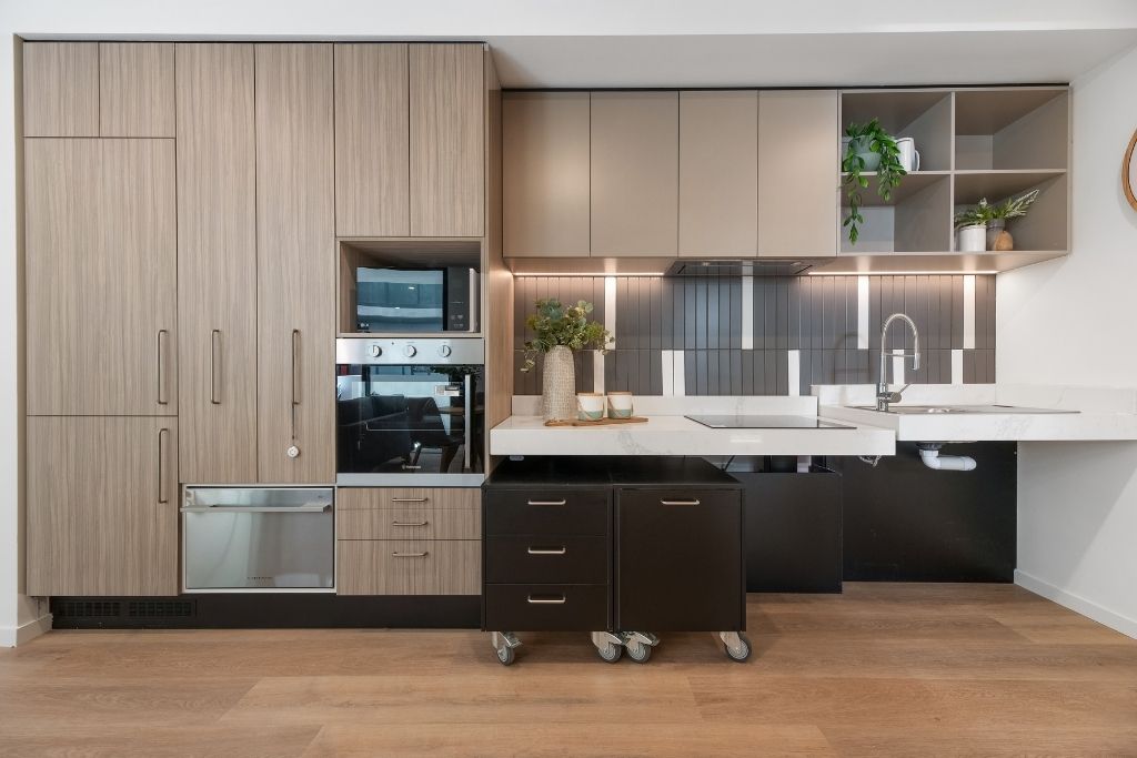 Accessible kitchen inside the Essendon SDA apartment, showing height-adjustable benches on castors, integrated dishwasher, wall oven, cooktop and stone benchtops.