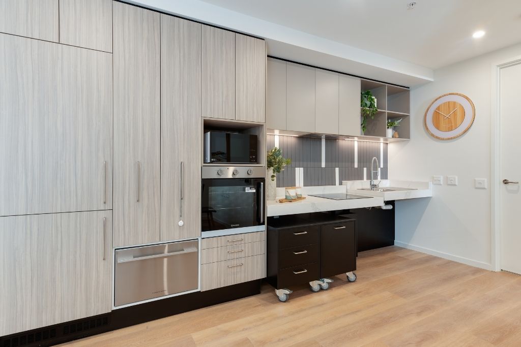 Accessible kitchen inside the Essendon SDA apartment, showing timber-look cabinetry, height-adjustable benches, wall oven, microwave and integrated dishwasher.