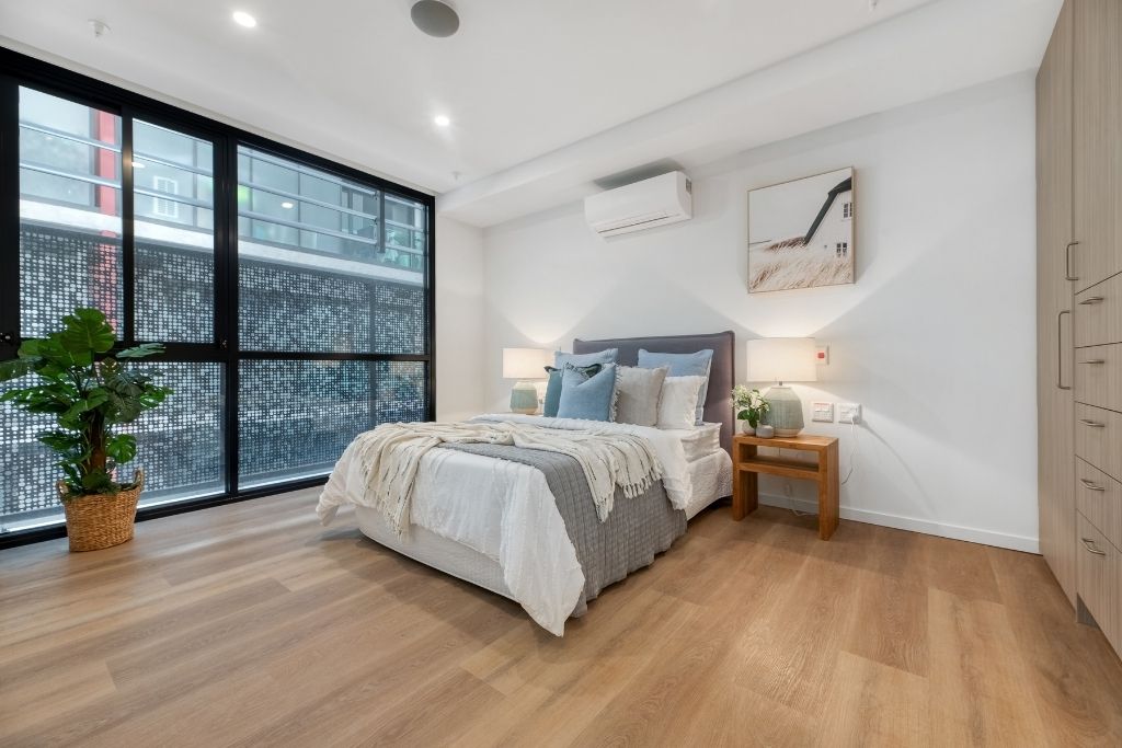 Bedroom inside the Essendon SDA apartment, showing timber-look flooring, split system air conditioning, built-in wardrobe and floor-to-ceiling windows.