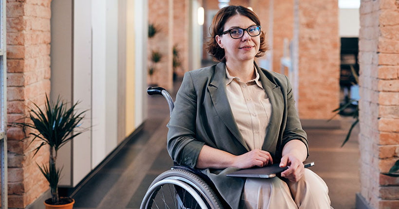 A woman holding a laptop, sitting in a wheelchair in an open plan office